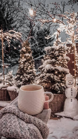 在雪地庭園中，手拿著一個用針織毛衣包裹的馬克杯，旁邊有雪人和燈光。 — 冬天 壁紙