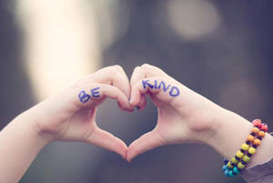 兩隻手形成心形，手掌上寫有「BE KIND」，一隻手腕上戴著彩色手鐲。