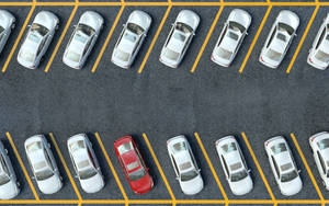 Rows of silver cars with one red car in a parking lot viewed from above.