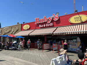 人群聚集在陽光明媚的海濱長廊上的Ruby's Bar & Grill，有條紋遮陽篷和食物標誌。