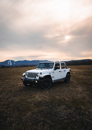 白色Jeep Wrangler停放在田野中，背景有山脈和夕陽。
