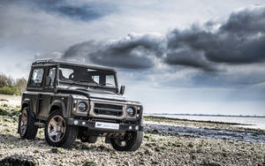 啞光黑色 Land Rover Defender 停在岩石海灘上，天空風暴來臨