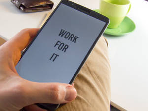 手拿著手機，旁邊有咖啡杯和錢包，顯示 'WORK FOR IT' 訊息。