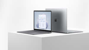 兩部銀色 Microsoft Surface 筆記型電腦，一部打開一部關閉，放在白色底座上。 — 美學筆電 壁紙
