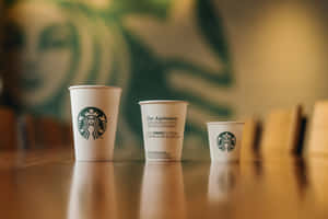 三個Starbucks咖啡杯排列在木製櫃檯上，背景是綠色標誌牆。 — 早晨咖啡 壁紙