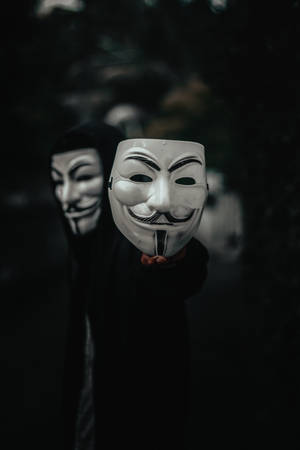 穿著黑色連帽衫的人在黑暗環境中手持白色蓋伊·福克斯面具。