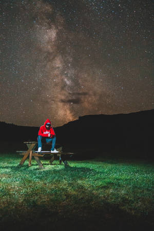 穿紅色連帽衫的人坐在野餐桌旁，星空下銀河拱橋