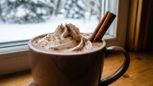 一杯熱朱古力配忌廉和肉桂棒，放在雪景窗邊。