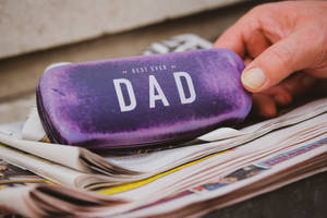 手拎住一個標有 'BEST EVER DAD' 嘅紫色眼鏡盒喺報紙上面。