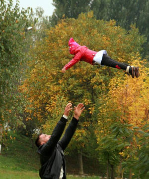 一名男子在秋天的公園裡將穿著粉紅色外套的小孩拋向空中。