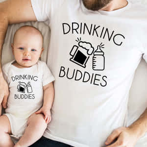 嬰兒和爸爸穿著相配的「drinking buddies」T恤在床上依偎。 — 配對 壁紙