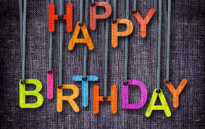 用色彩繽紛的布料製成的 happy birthday 字母，掛在繩子上，背景為深色紋理。