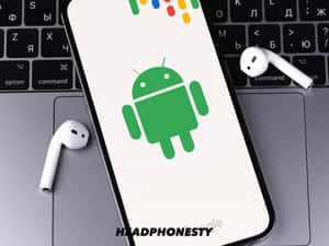 智能手機屏幕上顯示Android標誌，旁邊有手提電腦和白色耳機。 — 安卓 壁紙
