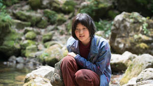 年輕女孩坐在森林中的溪流旁，穿著藍色外套和紅色褲子。