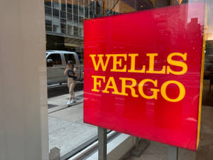 一個帶有黃色字樣的紅色 Wells Fargo 標誌反射出一個女人在城市人行道上行走。