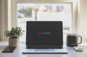 螢幕顯示「JUST START.」的筆記型電腦，旁邊有植物、杯子和電話在窗台上。
