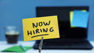 寫著 now hiring 的黃色便利貼夾在桌上的手提電腦上，旁邊有咖啡。