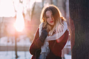 身穿冬季外套和圍巾的女性靠在雪地公園的樹上，陽光照射。