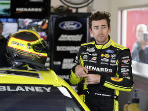 NASCAR車手Ryan Blaney身穿賽車裝備，站在車庫內他的黃色賽車旁。