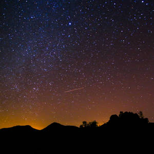 一顆流星劃過繁星點點的夜空，上方是黑暗的山脈輪廓。 — 月色夜空 壁紙