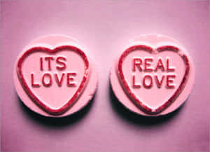 兩粒粉紅色心形糖果，帶有紅色閃粉文字，上面寫著「ITS LOVE」和「REAL LOVE」。