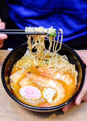 雙手用筷子從一碗蝦和魚餅湯中夾起拉麵。 — 美食 壁紙