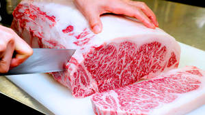 手在白色砧板上用刀切一大塊大理石紋烤肉。