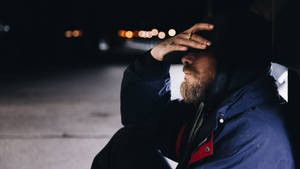 一個留鬍鬚的男人穿著連帽外套，夜晚獨自坐在街上，背後有模糊的燈光。