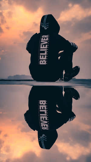 穿著 believer 連帽衫的人坐在水邊，帶有日落倒影的剪影。
