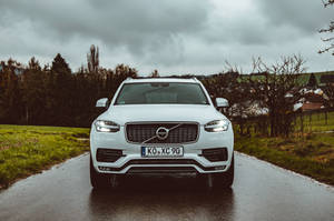 白色Volvo XC90 SUV在潮濕的鄉村道路上，多雲天空和綠色田野。