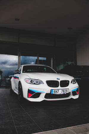 白色BMW M2跑車，帶有藍色和紅色點綴，停放在室內靠近玻璃門處，顯示多雲天空。