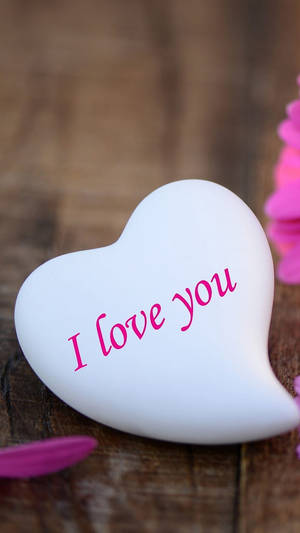 白色心形，上面有粉紅色字句寫住 I love you，放在木質表面上，伴有粉紅色花瓣。
