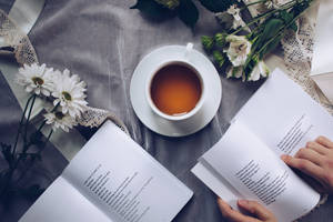 兩隻手在灰色布上閱讀打開的書，旁邊有一杯茶和白色花朵。