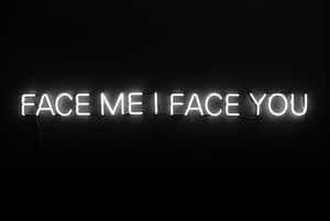 一個白色霓虹燈牌，上面寫著 face me i face you，掛在黑色牆上。