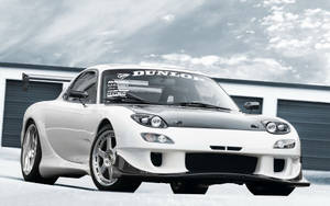 白色萬事得RX-7跑車，帶有黑色引擎蓋和賽車貼紙，停泊在車庫前。