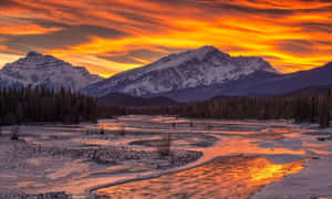 白雪覆蓋的山脈和河流在充滿活力的橙色日落天空下閃耀。 — 冬日美學 壁紙
