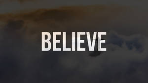 在黑暗的多雲天空背景下，粗體白色文字寫著「BELIEVE」，並帶有模糊的十字架輪廓。