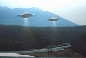 兩個UFO盤旋在一條有森林和白雪皚皚山峰的山路上。