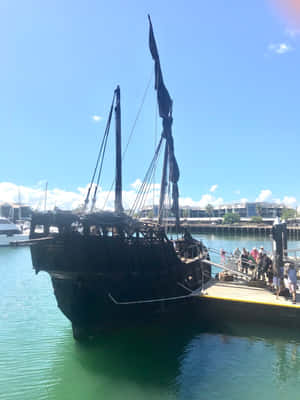 深色海盜船停泊在碼頭，遊客在棧橋上，藍天白雲。