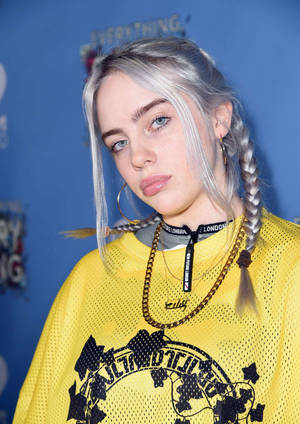 Billie Eilish 身穿黃色網眼上衣，梳著銀色辮子，戴著圓環耳環。