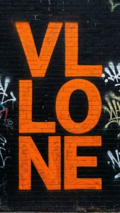 橙色塗鴉字母在黑色磚牆上拼寫 VLONE，帶有白色標籤。 — Vlone 壁紙