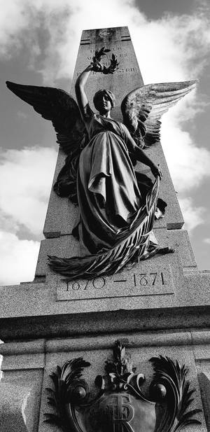 帶翼天使雕像矗立在歷史石製紀念碑上，日期為1870-1871年，天空多雲。
