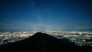 黑暗的山峰俯瞰著夜晚燈火通明的城市，星空下銀河清晰可見