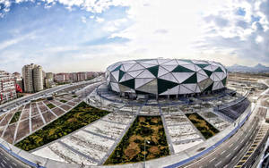 一個現代幾何圓頂體育場，在多雲的天空下被道路和城市建築環繞。 — 2560x1600 壁紙