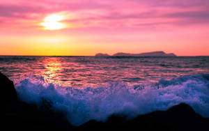 粉紅色和橙色的夕陽映照在海浪上，海浪拍打著深色岩石，遠處有島嶼。 — 美麗的日落動畫 壁紙