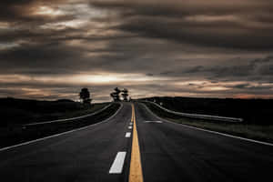 黃昏時分空無一人的蜿蜒道路，有烏雲和樹木剪影。 — 道路 壁紙