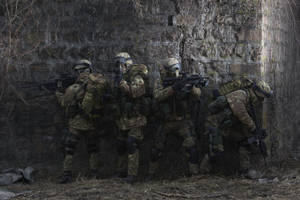 四名身穿迷彩裝備的武裝士兵蹲在石牆後面，處於戰鬥姿態。