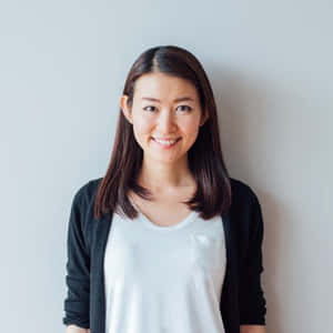 一位棕色頭髮的女性，身穿白色襯衫和黑色開襟毛衣，在灰色牆前微笑。