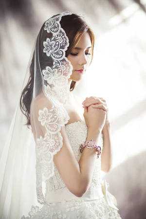 身穿白色蕾絲婚紗的新娘在柔和窗光下合十祈禱。