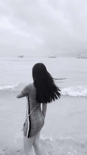 身穿格子裙的女性站在海邊，背景有波浪和船隻，霧氣朦朧。